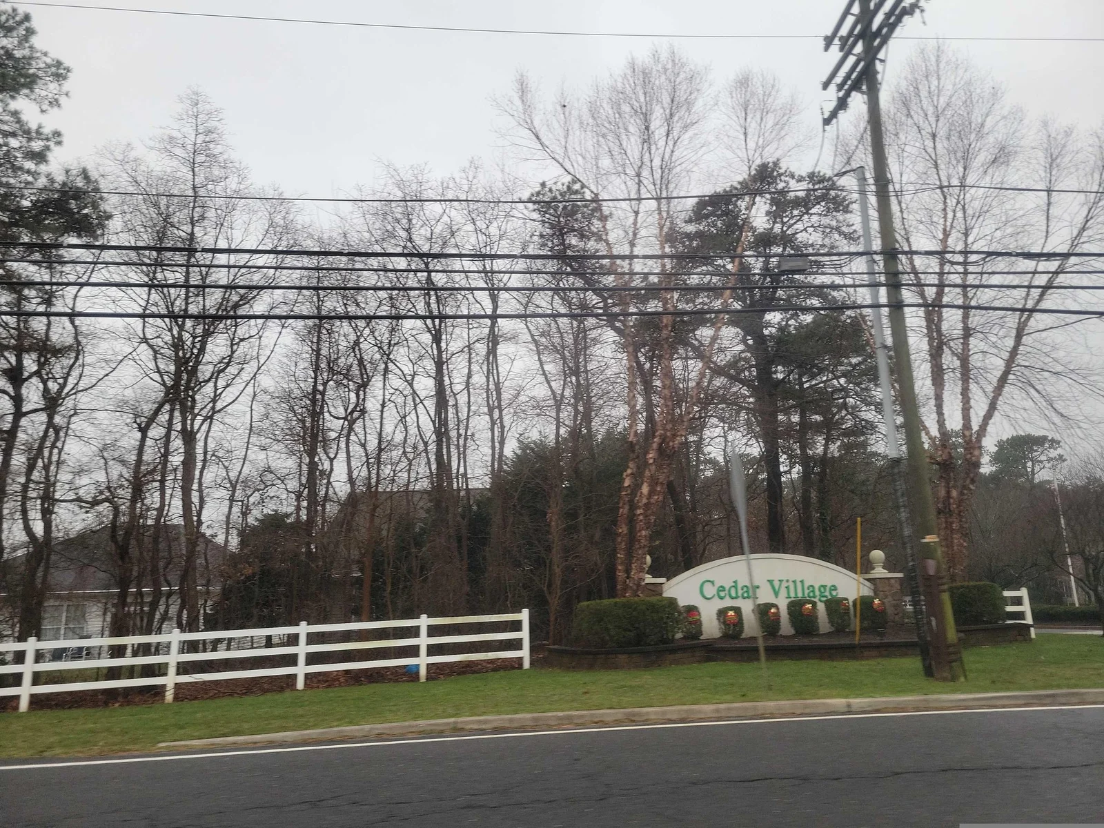 Community sign and white fence under tall trees beside the road