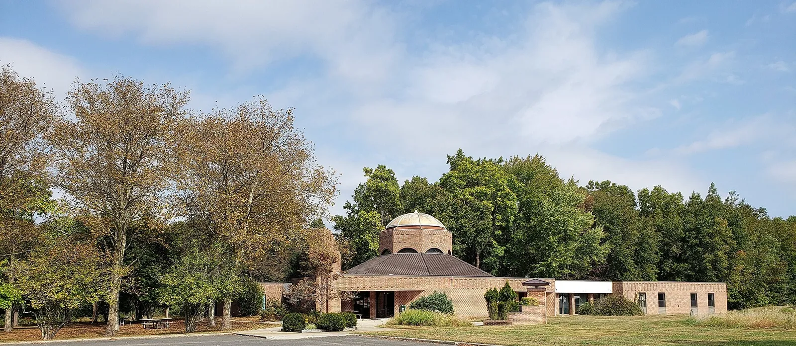 A low brick building with a domed rotunda set among tall trees under a soft blue sky