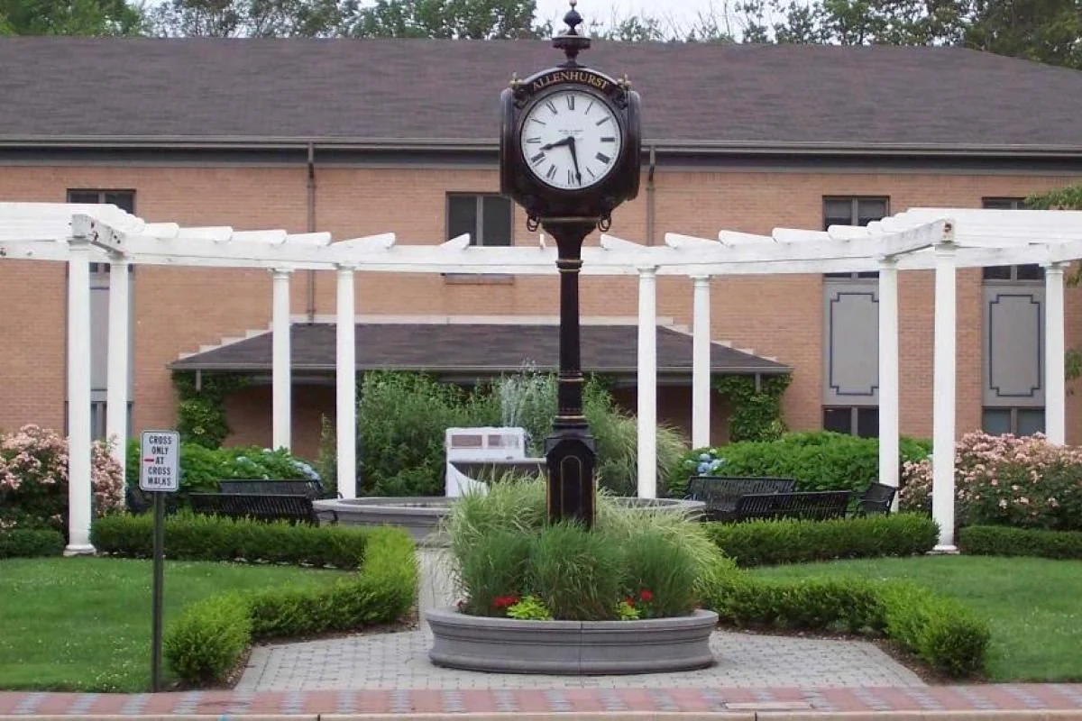 A black post clock in a round planter framed by a white colonnade and flower beds in Allenhurst