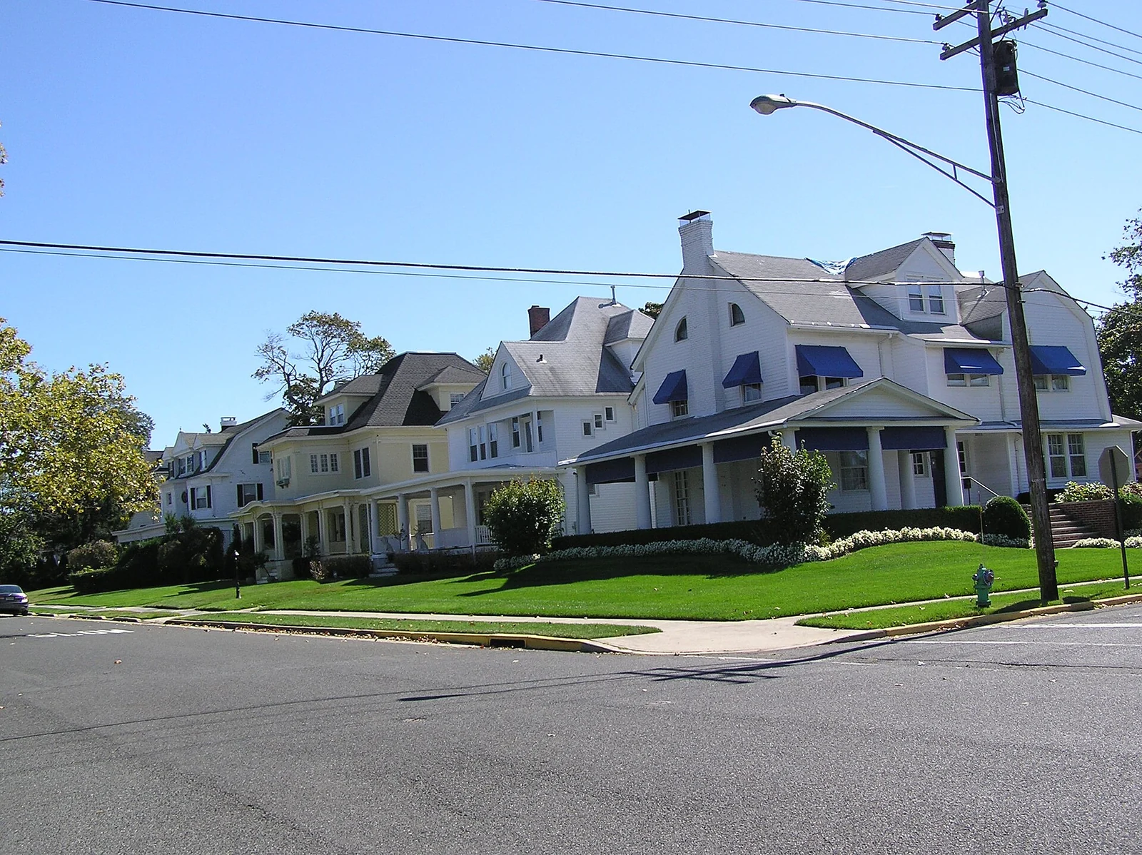 A row of large white historic homes with porches and blue awnings across a green corner lawn in Allenhurst