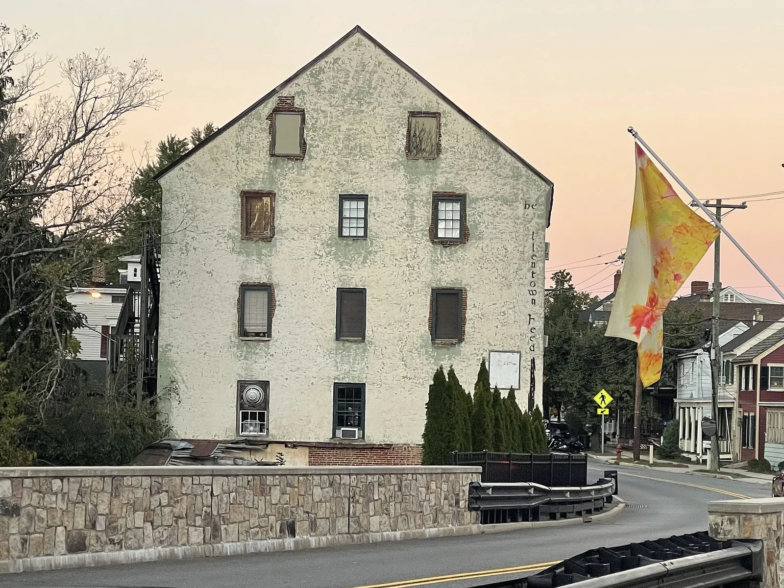 Tall historic stucco mill building beside a stone bridge wall under a pink evening sky