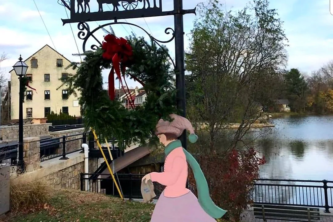 A holiday wreath hanging from a wrought-iron horse-and-carriage sign and a painted caroler figure beside the millpond at Pete Sensi Park, the mill behind