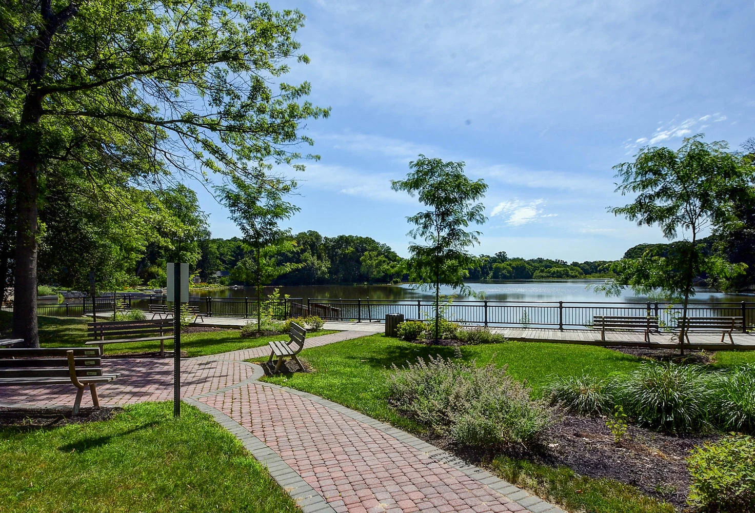 A brick walkway with benches leading through Pete Sensi Park to Conines Millpond on a sunny day