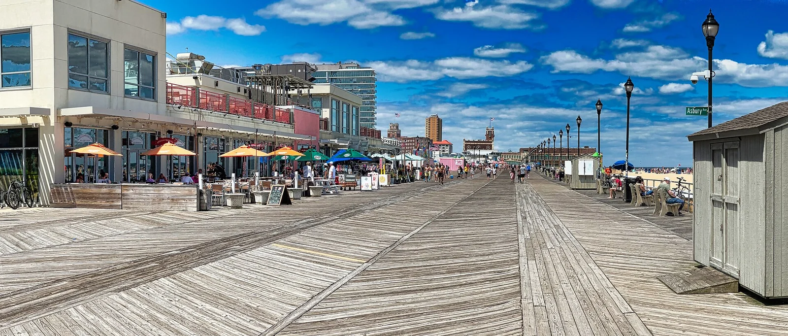 Wide view of the Asbury Park boardwalk with outdoor restaurant seating and umbrellas on the left under a blue summer sky