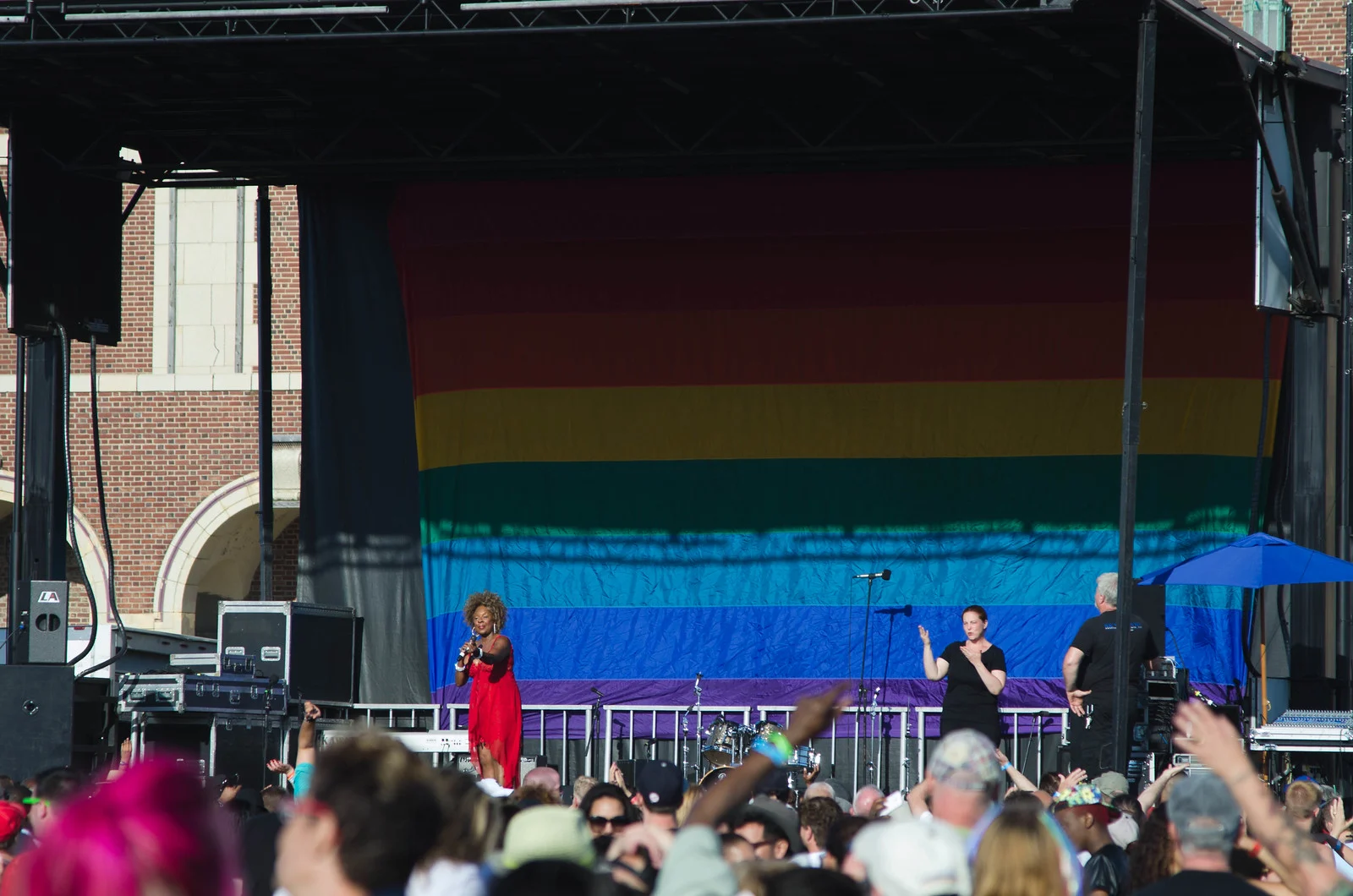 A singer in red performing on an outdoor stage hung with a giant rainbow flag, a crowd cheering in front