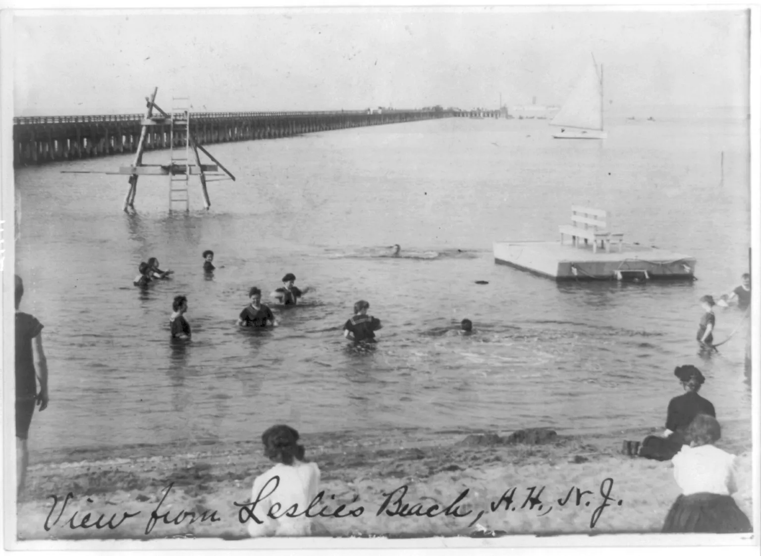 Black-and-white photo of bathers wading in the bay beside a diving platform, with a long pier stretching out behind them