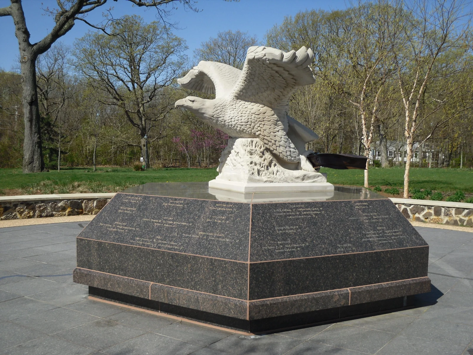 White stone eagle holding a steel beam atop a black granite base engraved with names, at Monmouth County's 9/11 Memorial in spring