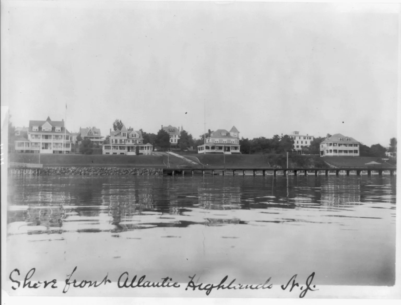 Black-and-white photo of large Victorian houses lining the waterfront above calm bay water