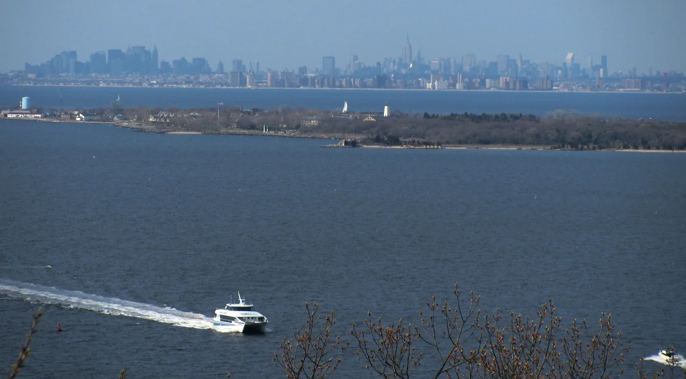 A ferry crossing the bay below the Atlantic Highlands hills, with Sandy Hook and the New York City skyline beyond