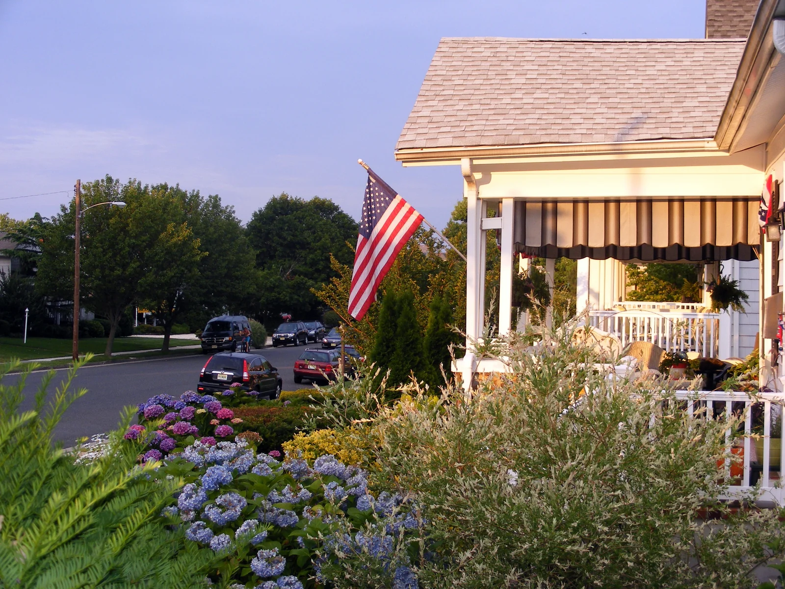 A white front porch with a striped awning and an American flag above blooming hydrangeas on Garfield Avenue, the street and trees beyond