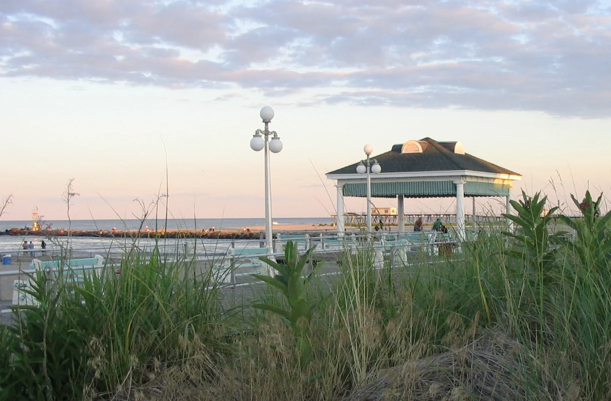 A green-roofed boardwalk pavilion and globe lampposts behind tall dune grass at the Shark River Inlet end of the boardwalk at dusk