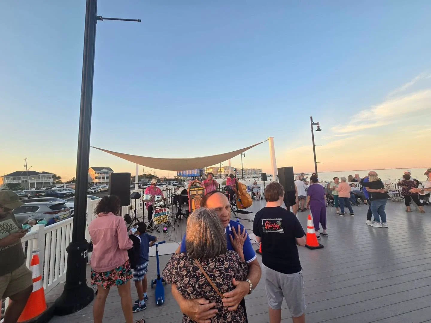 Couples dancing on the Barnegat dock deck while a band plays under a shade sail, with the bay and a pink sunset sky behind