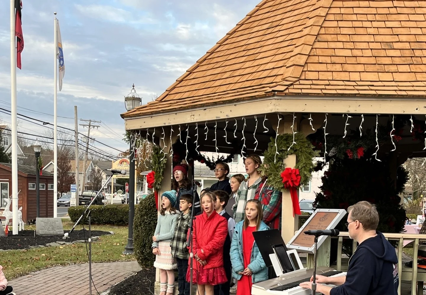 A children's choir in red and holiday colors singing at the wreath-hung gazebo while a pianist plays a keyboard