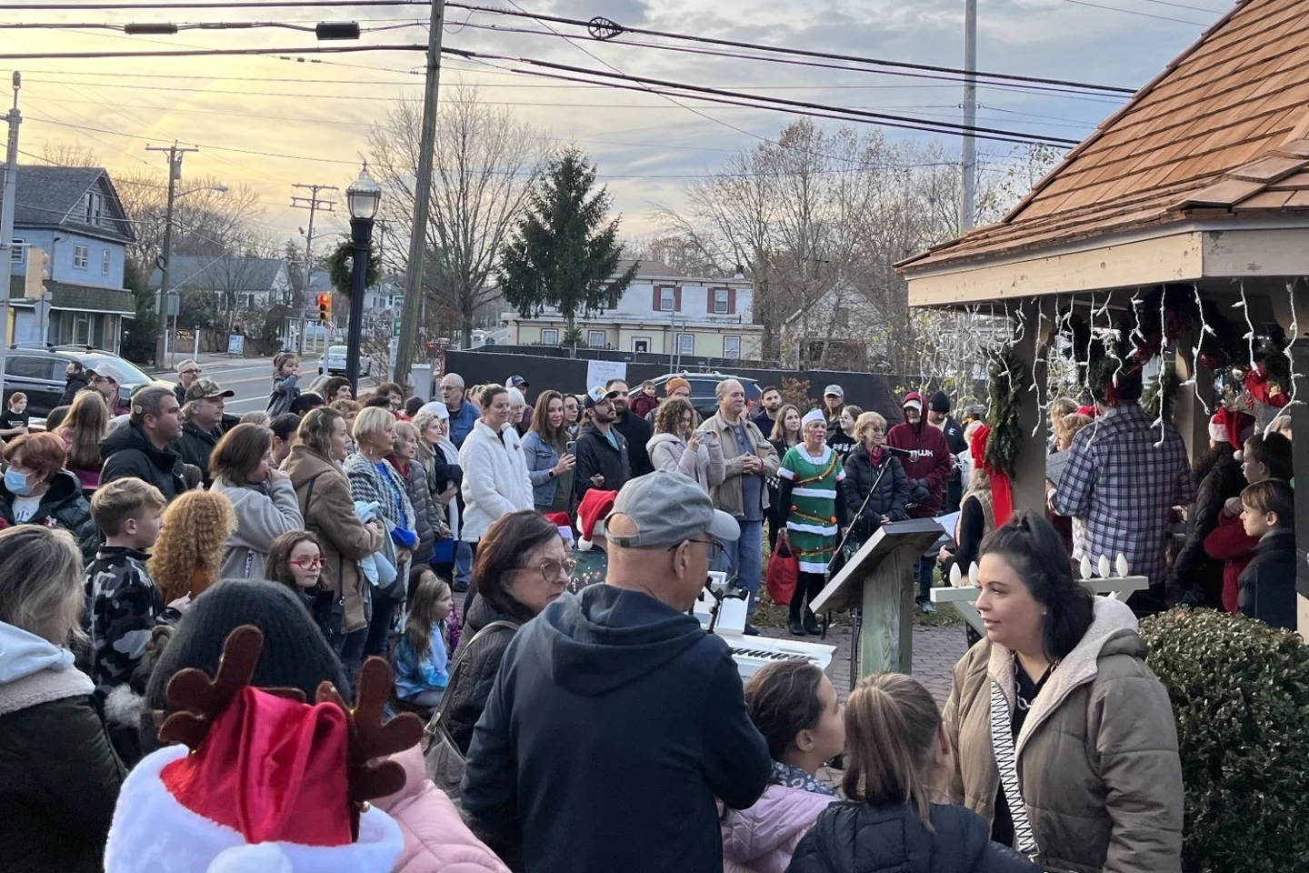 A crowd in winter coats and Santa hats gathered around the decorated township gazebo at dusk