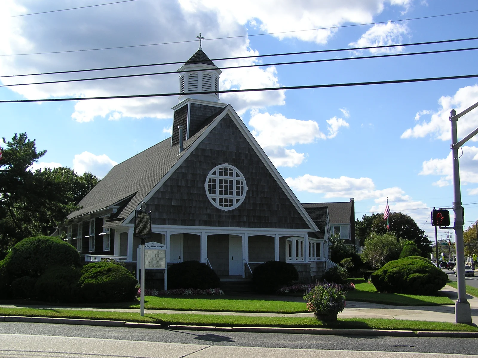The shingled Bay Head Chapel with a white porch, round window and cupola, behind trimmed shrubs