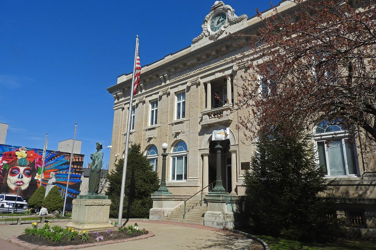 The ornate stone Belleville Town Hall with a flagpole and flower bed, and a colorful mural beside it