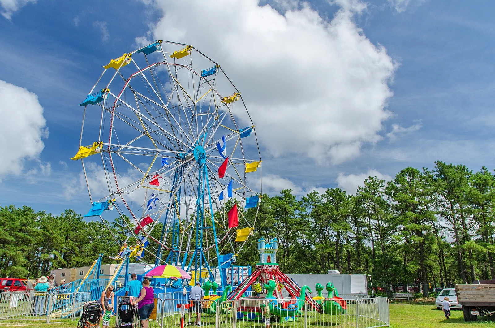 A Ferris wheel with colorful flags and kiddie rides under a blue sky, pine trees behind, at the Ocean County Fair