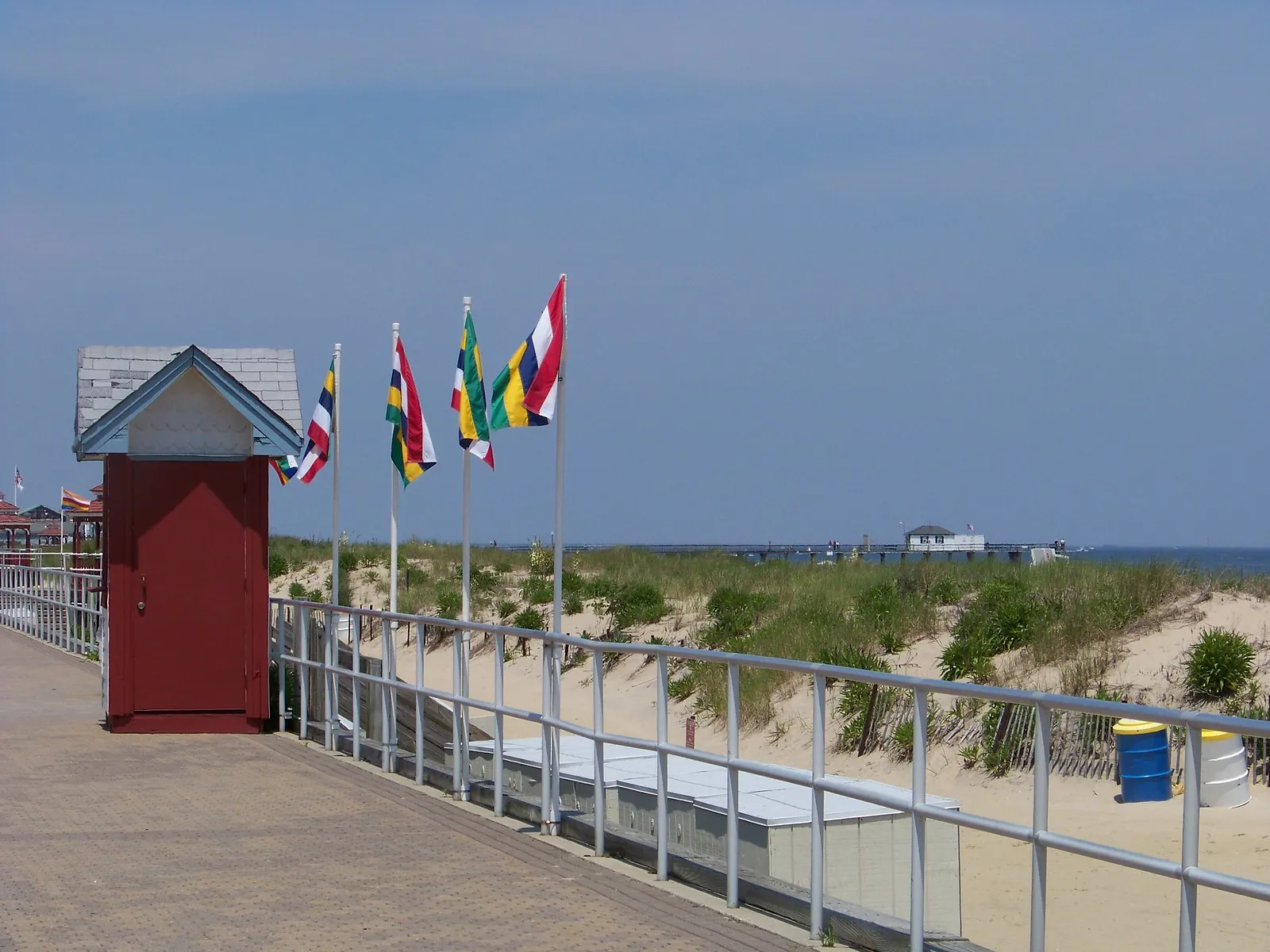 A row of colorful flags and a small red booth along the Bradley Beach boardwalk, with dune grass beside the railing