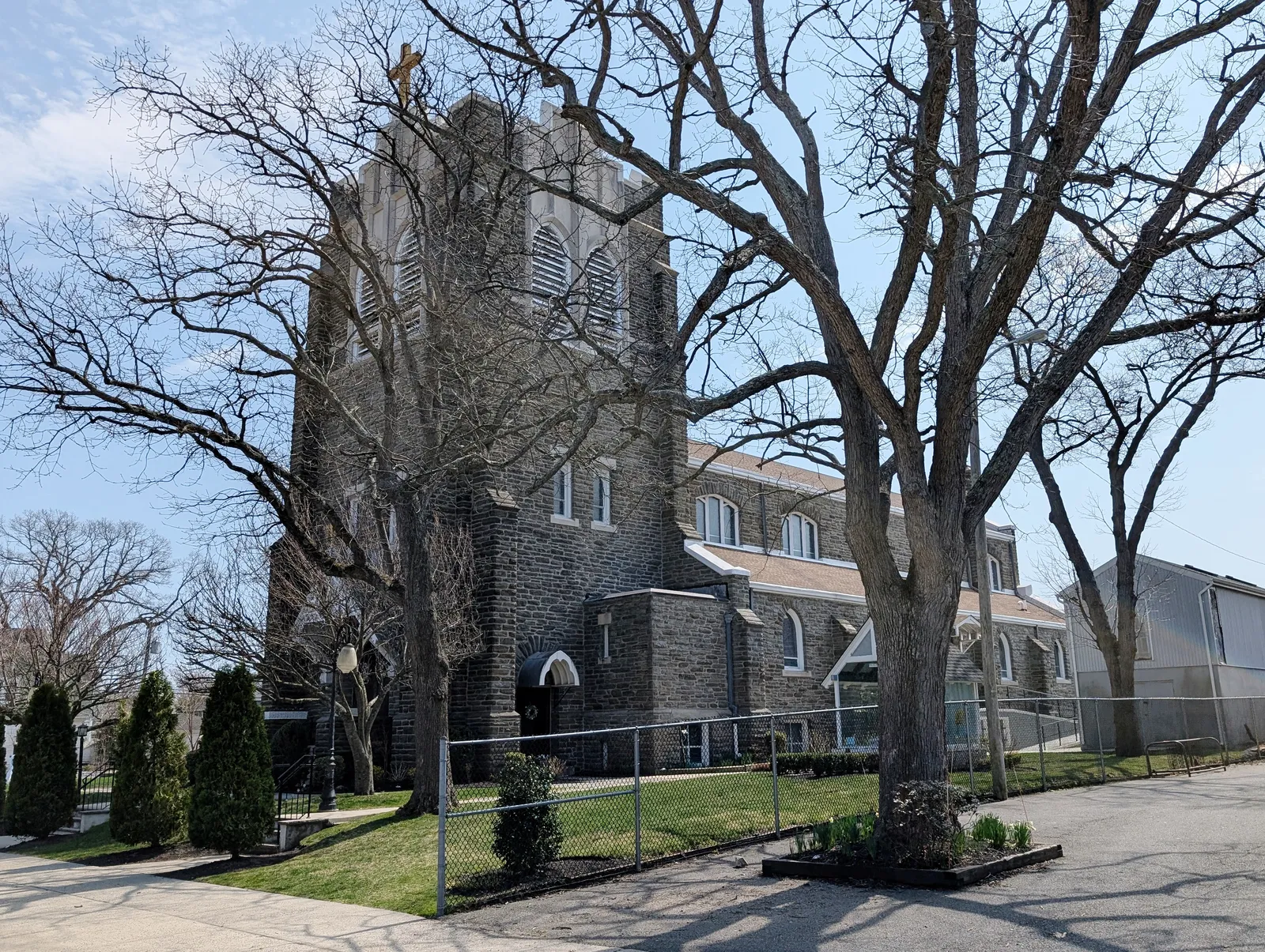 A gray stone church with a square bell tower behind tall bare trees under a blue sky
