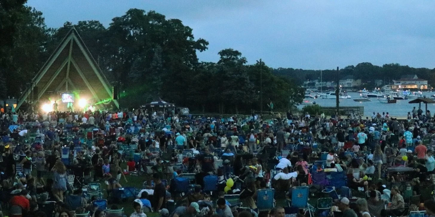 A packed lawn at dusk in front of the lit Windward Beach Park bandshell, boats on the river behind