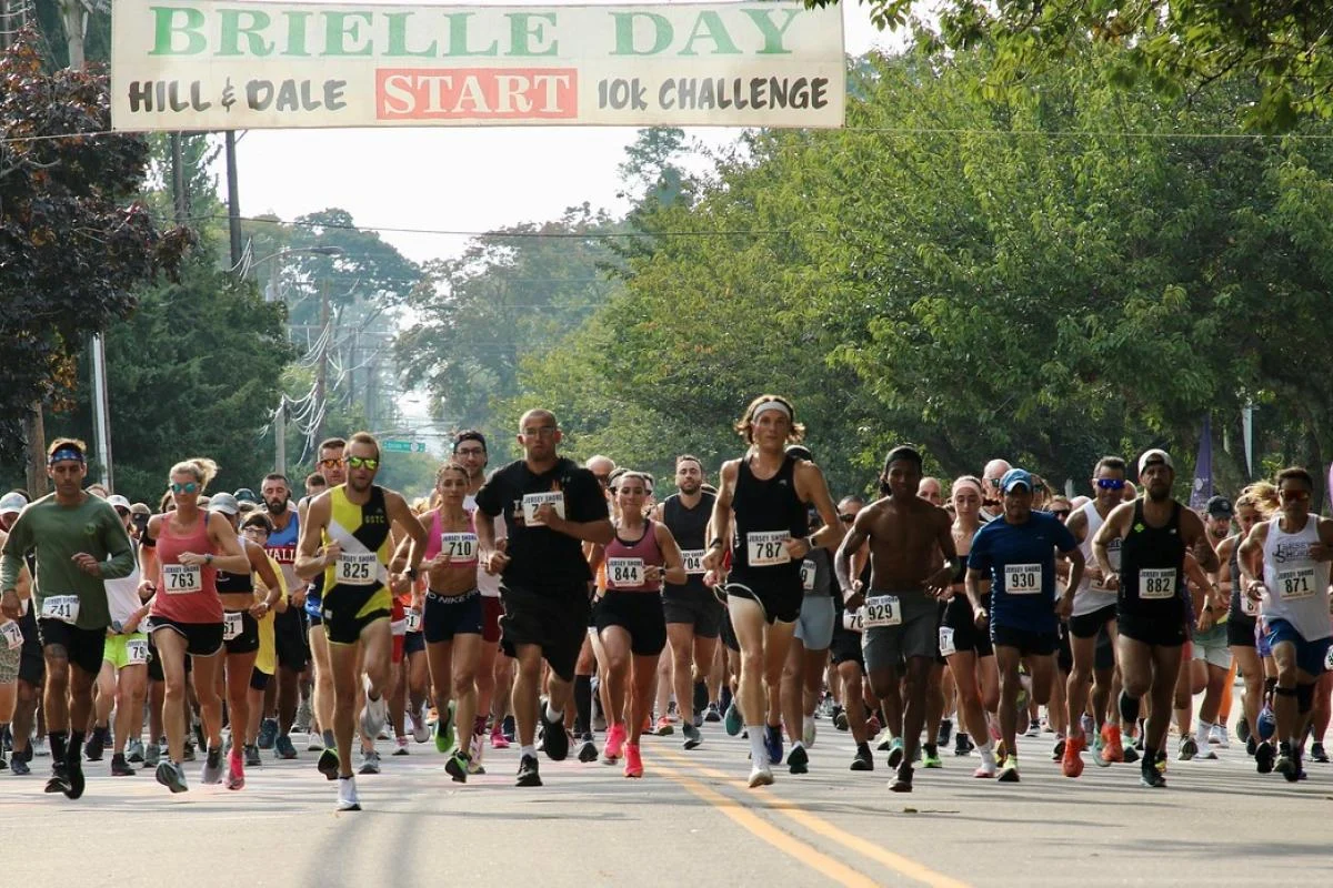 A pack of runners leaving the start line under a Brielle Day Hill and Dale 10K Challenge banner on a tree-lined street