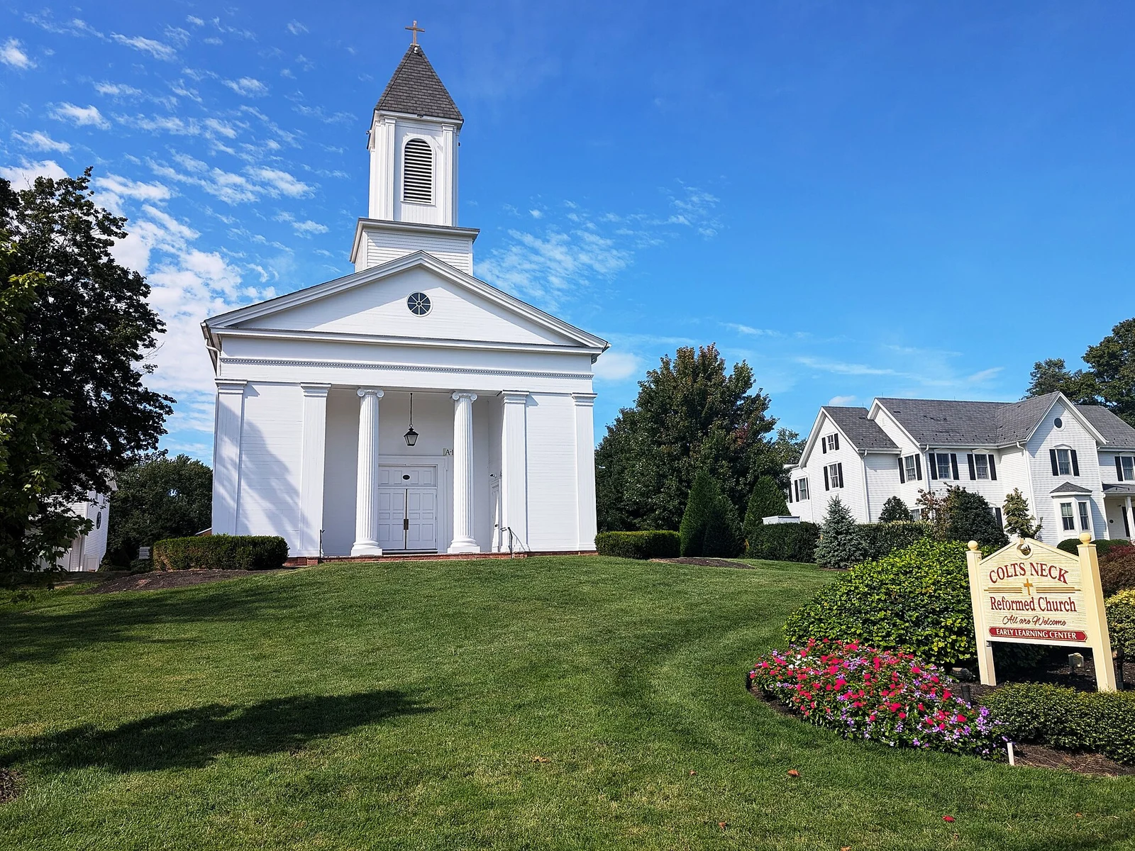 The white columned Colts Neck Reformed Church with its steeple, a green lawn and a flower bed by the sign
