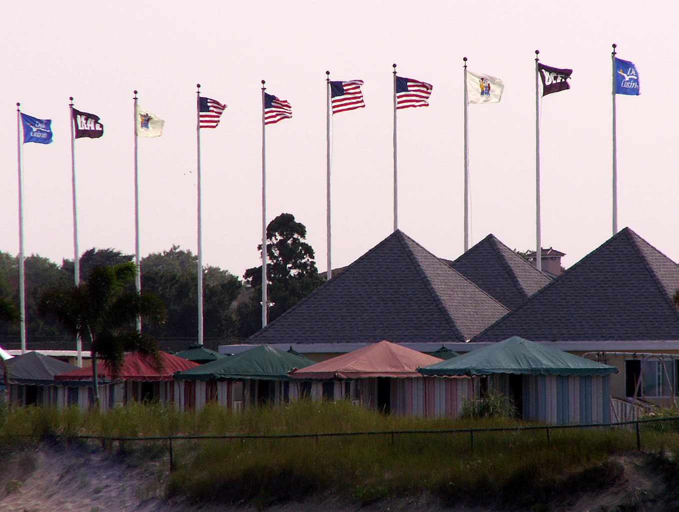 Flags flying on tall poles above peaked cabana roofs and striped tents at a Deal beach club
