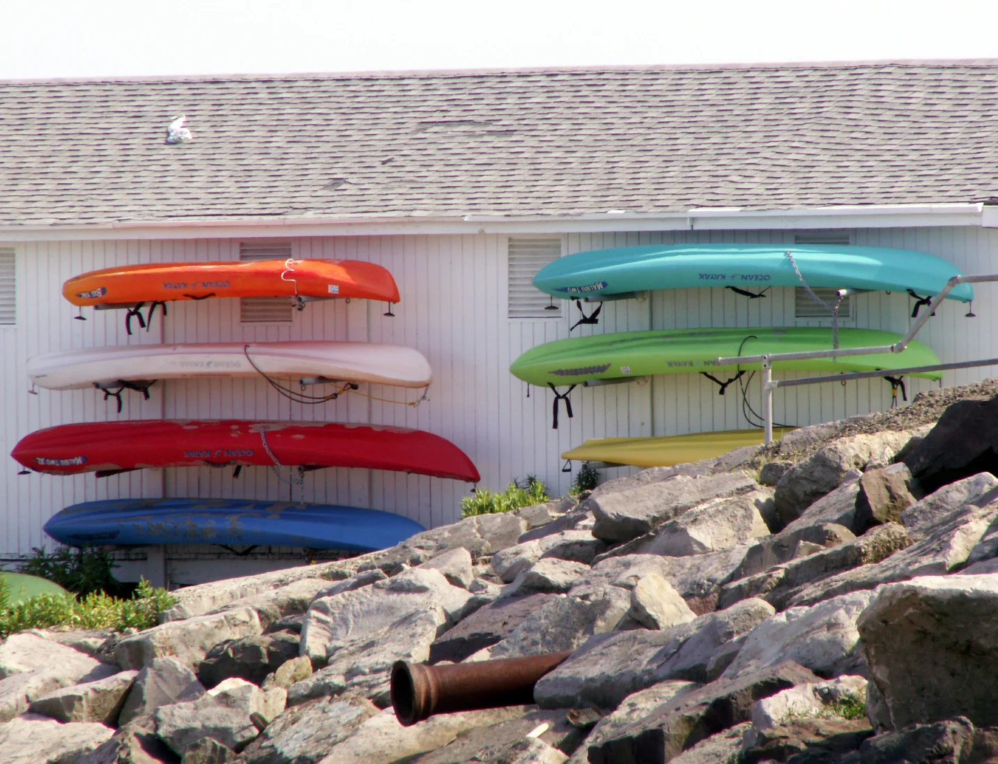 Orange, red, blue and green kayaks racked on a white beach club wall above rocks in Deal