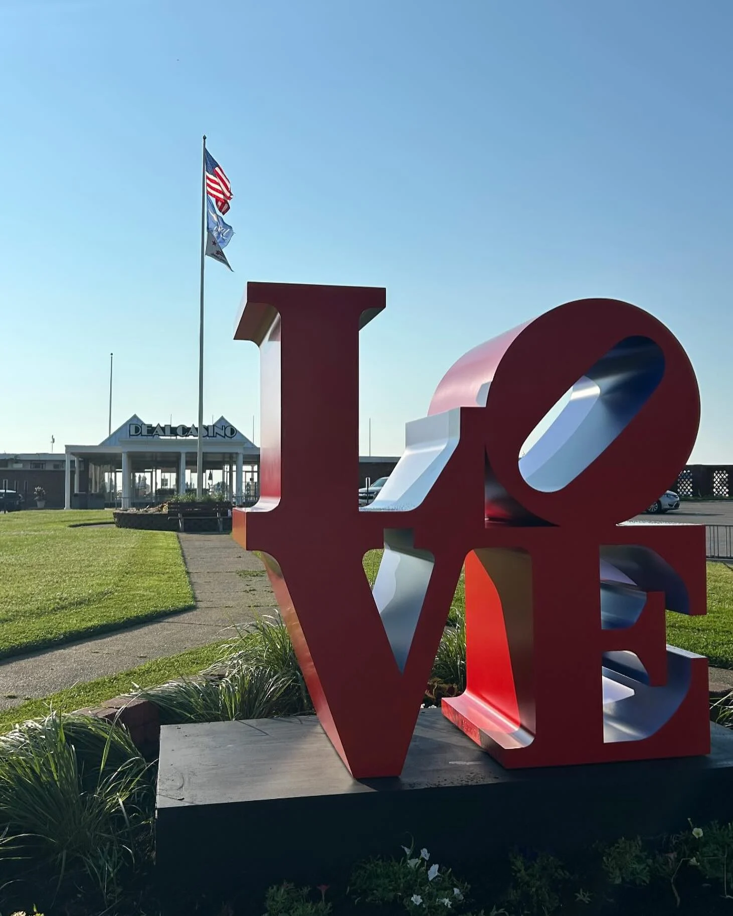A red LOVE letter sculpture on a planted base on the lawn in front of the Deal Casino Beach Club, with flags flying overhead