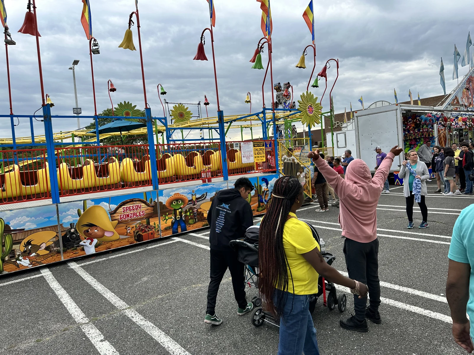 Visitors walking past a colorful kiddie roller coaster and game booths at a carnival in the Monmouth Mall parking lot