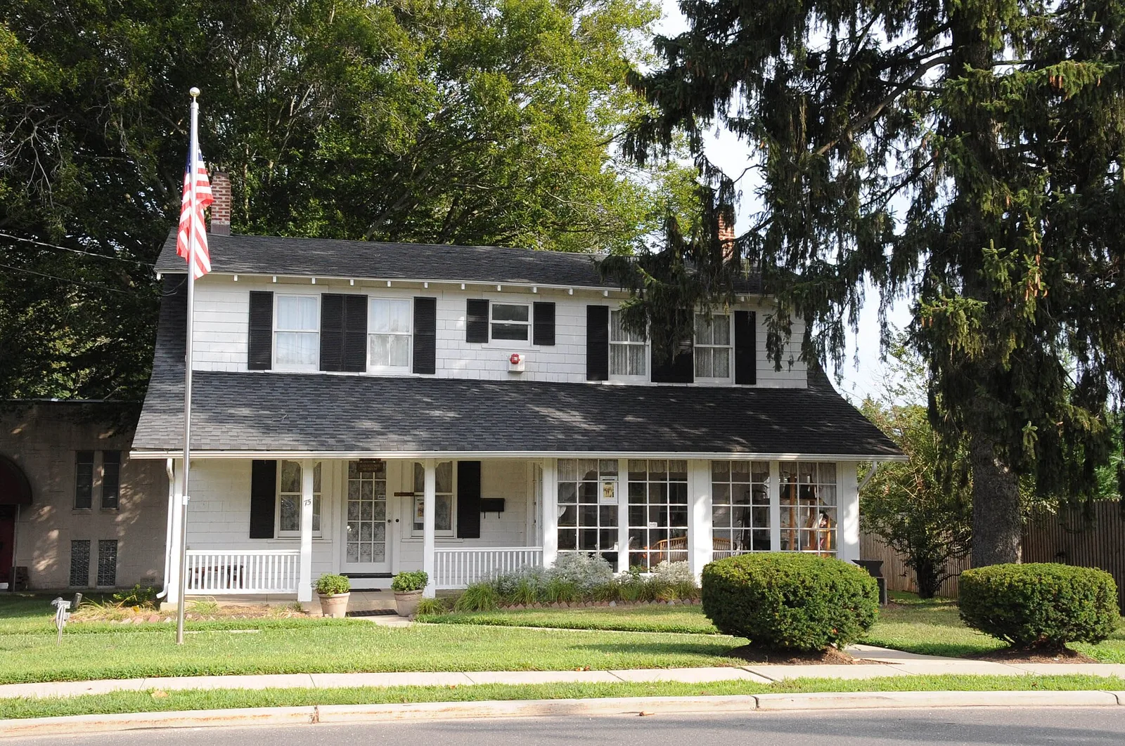 A white two-story colonial village house with black shutters and a flagpole on the front lawn