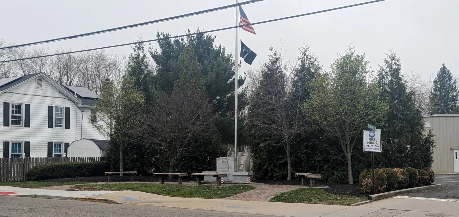 A small memorial garden with stone benches, a monument and a flagpole flying the American and POW-MIA flags, backed by evergreens