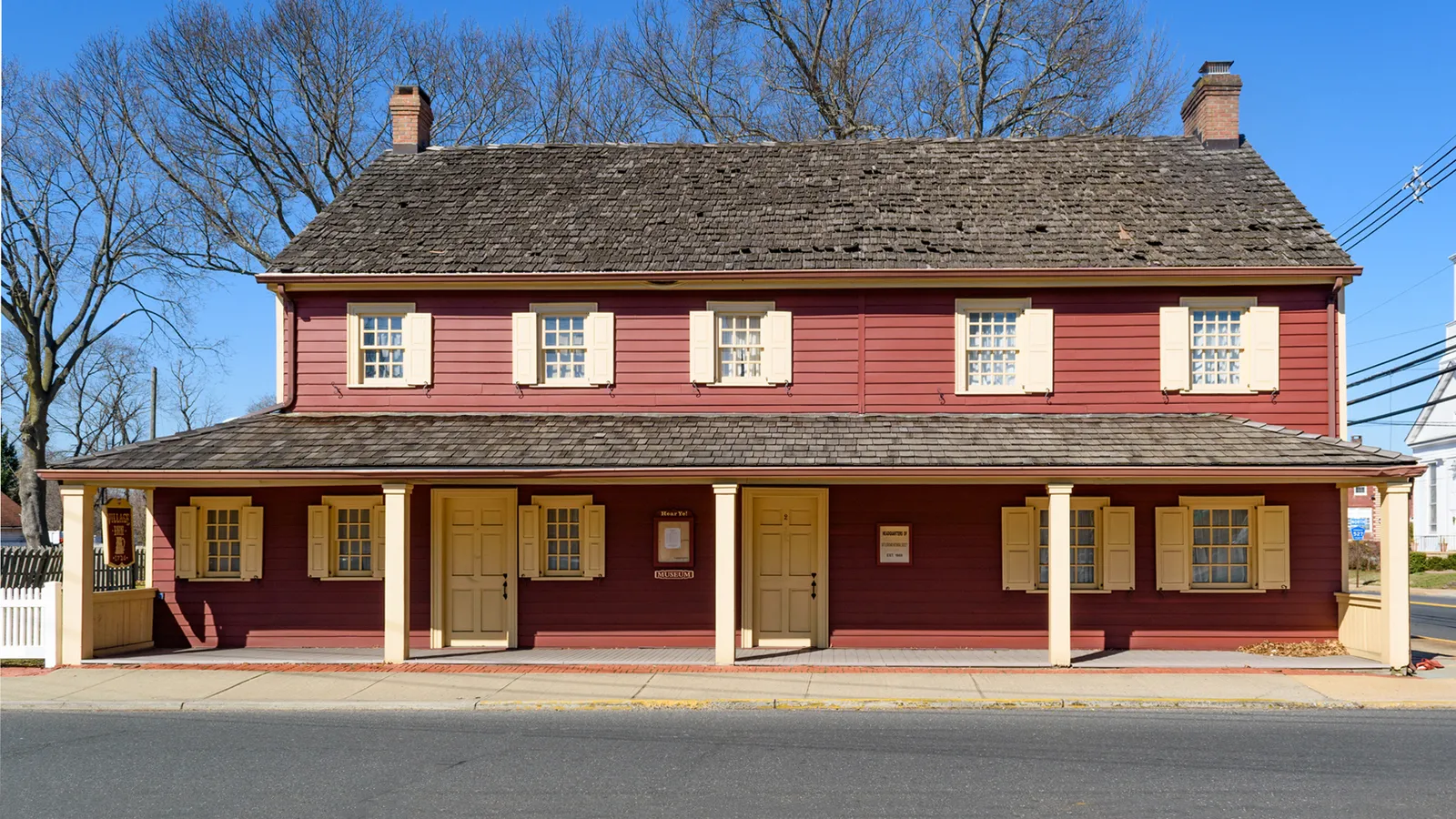 A red two-story colonial inn with cream shutters and a long front porch under a clear blue sky