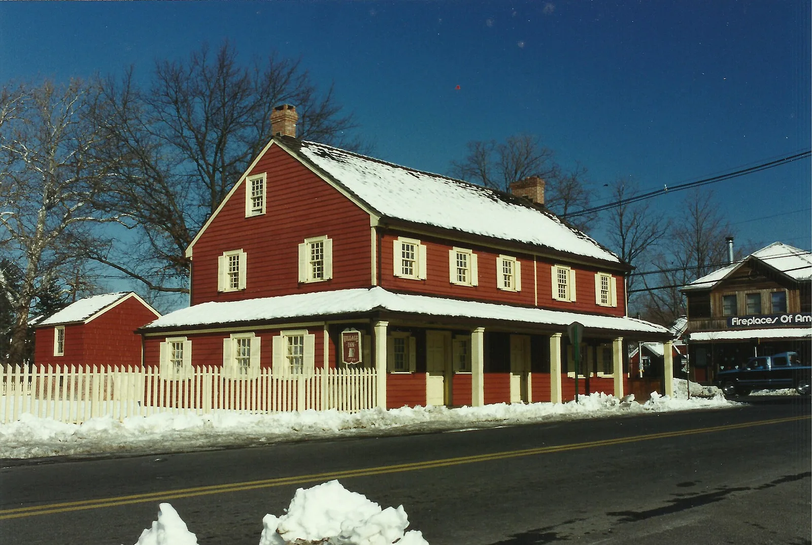 The red Village Inn in Englishtown after a snowfall, behind a white picket fence under a deep blue sky