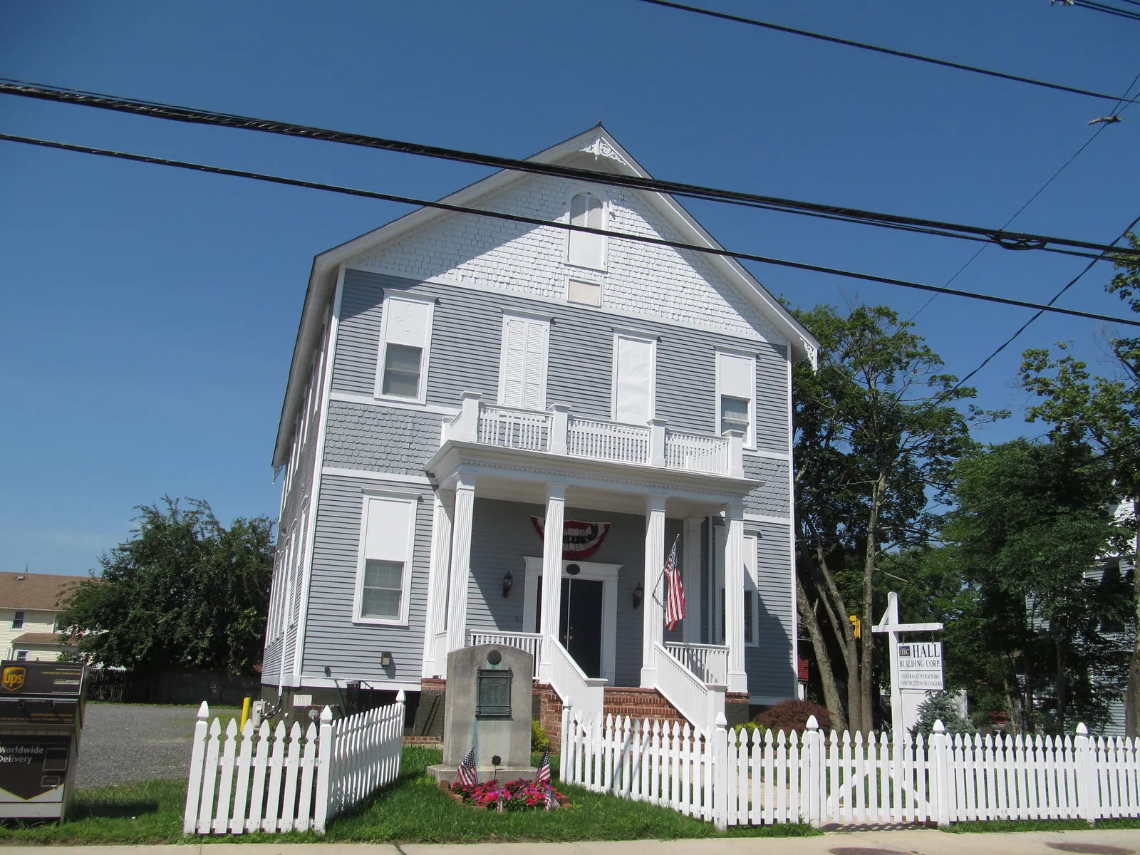 A tall clapboard building with a columned porch, a white picket fence and a stone memorial marker with flags in front