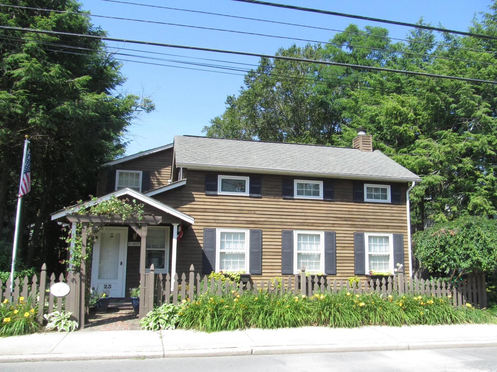 A brown-shingled two-story home with a vine-covered arbor, a picket fence and a bed of daylilies
