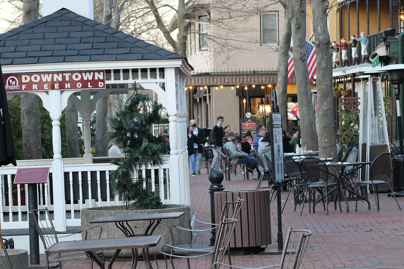 A white gazebo with a Downtown Freehold sign beside outdoor tables on a brick plaza