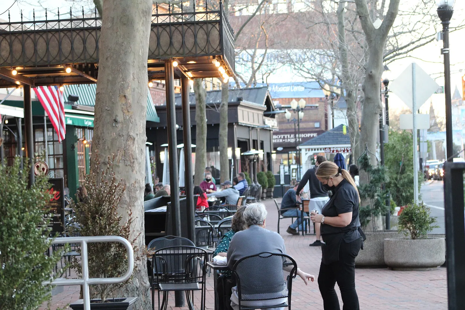 Diners at sidewalk tables under trees and string lights on a downtown street