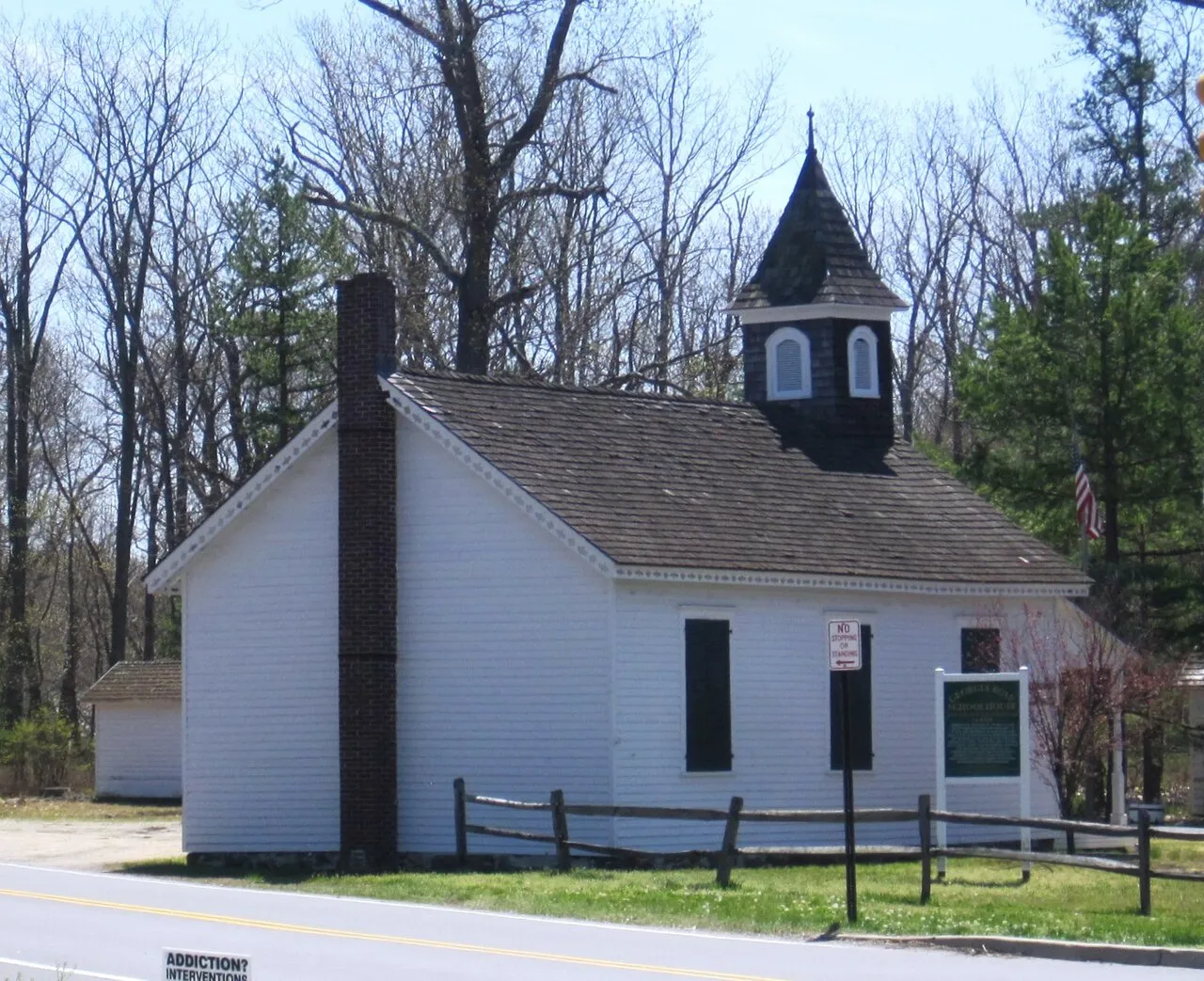 A small white one-room schoolhouse with a dark cupola and a brick chimney beside a wooded roadside
