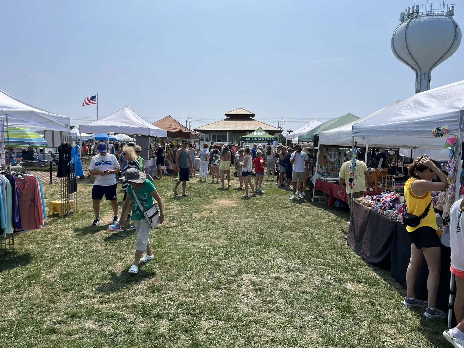 Shoppers browsing vendor tents on the lawn of Sunset Park, with the Harvey Cedars water tower and a gazebo roof behind