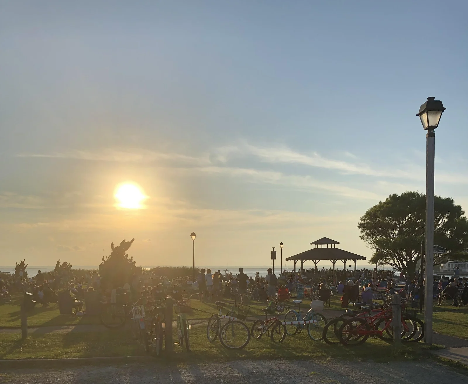 The sun setting over the bay behind a crowd on the lawn of Sunset Park, with a gazebo, lamp posts and bicycles parked in front