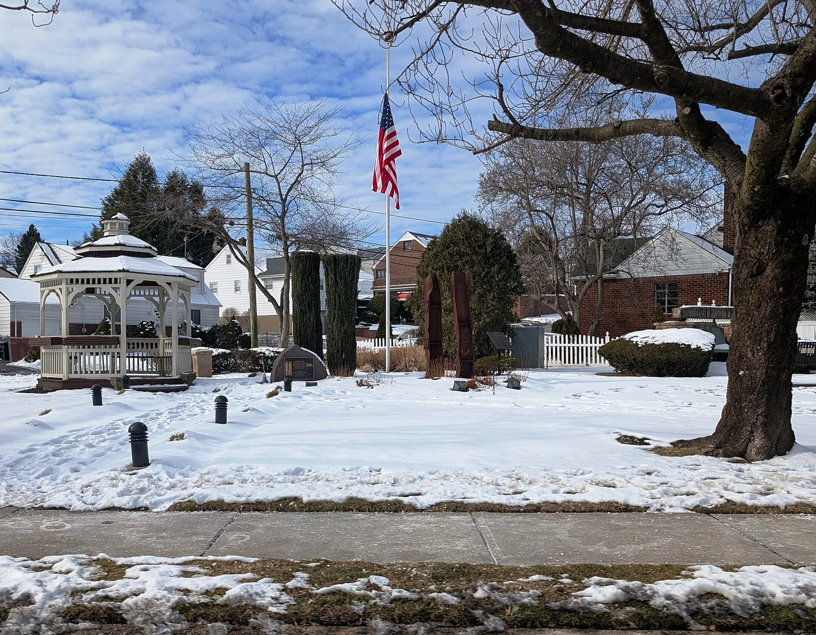 A white gazebo, flagpole and memorial on a snowy lawn surrounded by trees