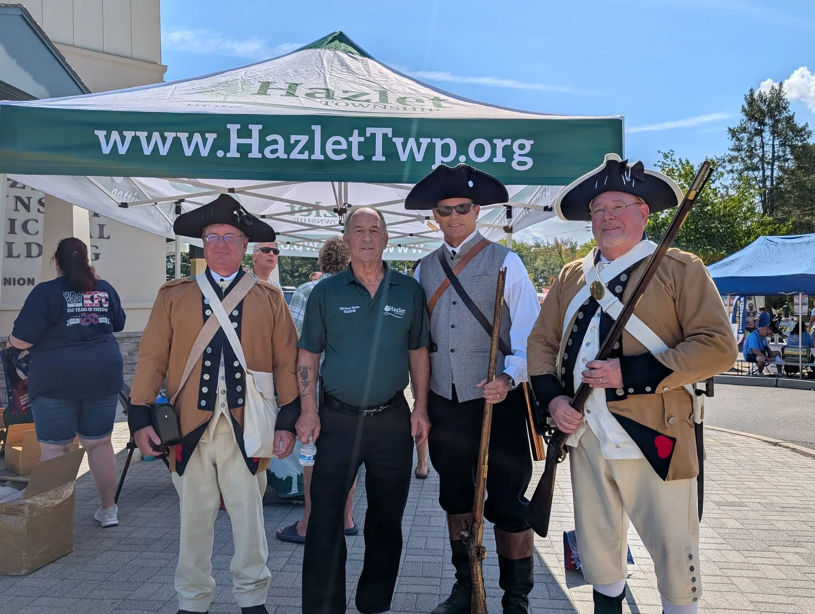 Revolutionary War reenactors in tricorn hats and colonial uniforms with muskets under a Hazlet Township tent at Hazlet Day