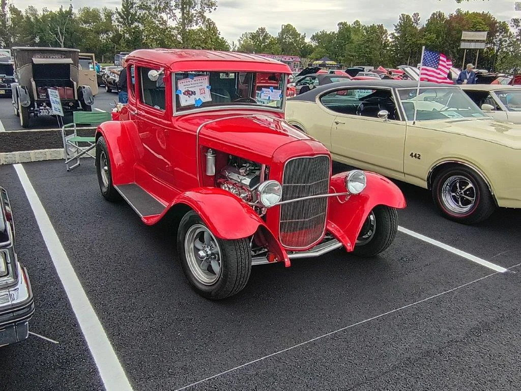 A bright red hot rod coupe with a chrome grille and flags flying over rows of classic cars