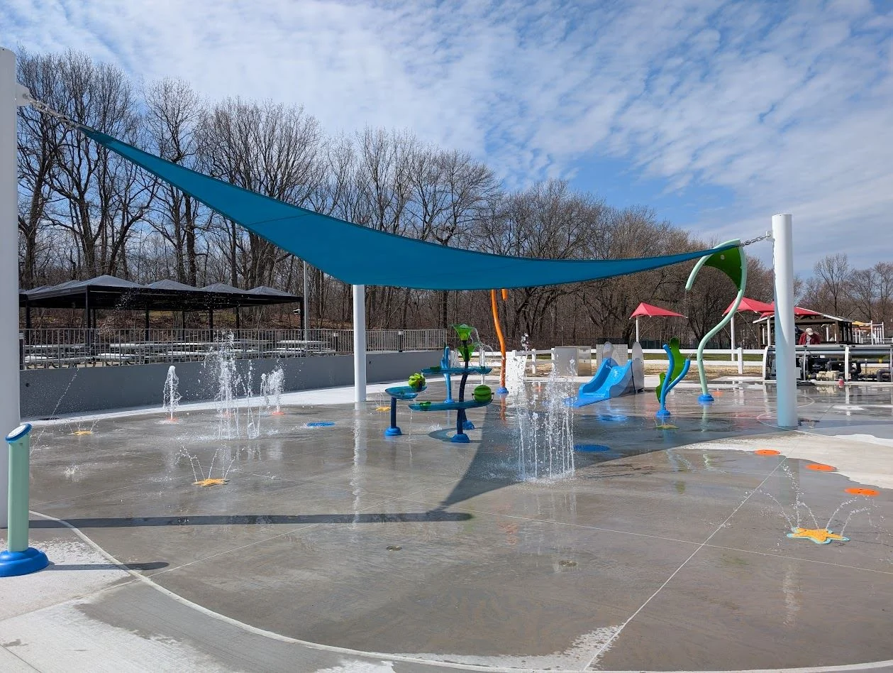 Water jets and colorful spray features under a blue shade sail at the Hazlet Swim & Tennis Club splash pad