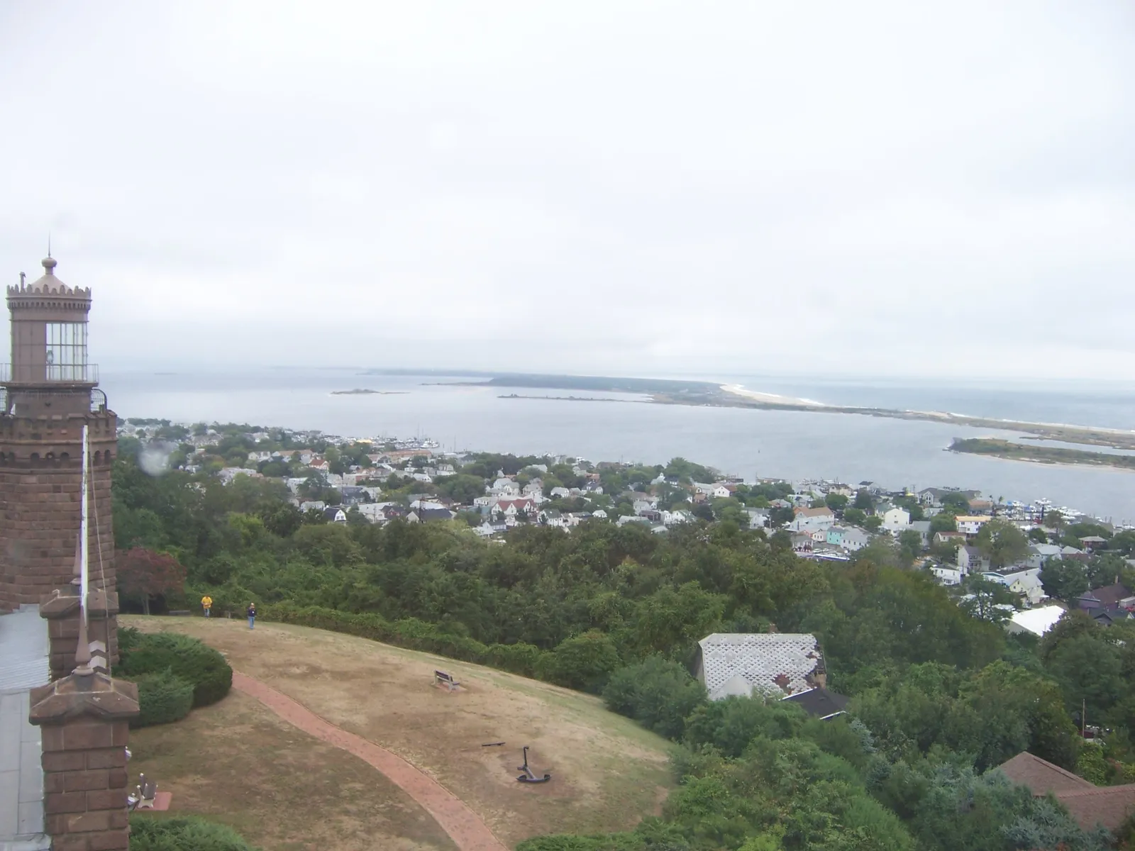 View from a lighthouse tower over a lawn, treetops and rooftops to the river and the long Sandy Hook peninsula