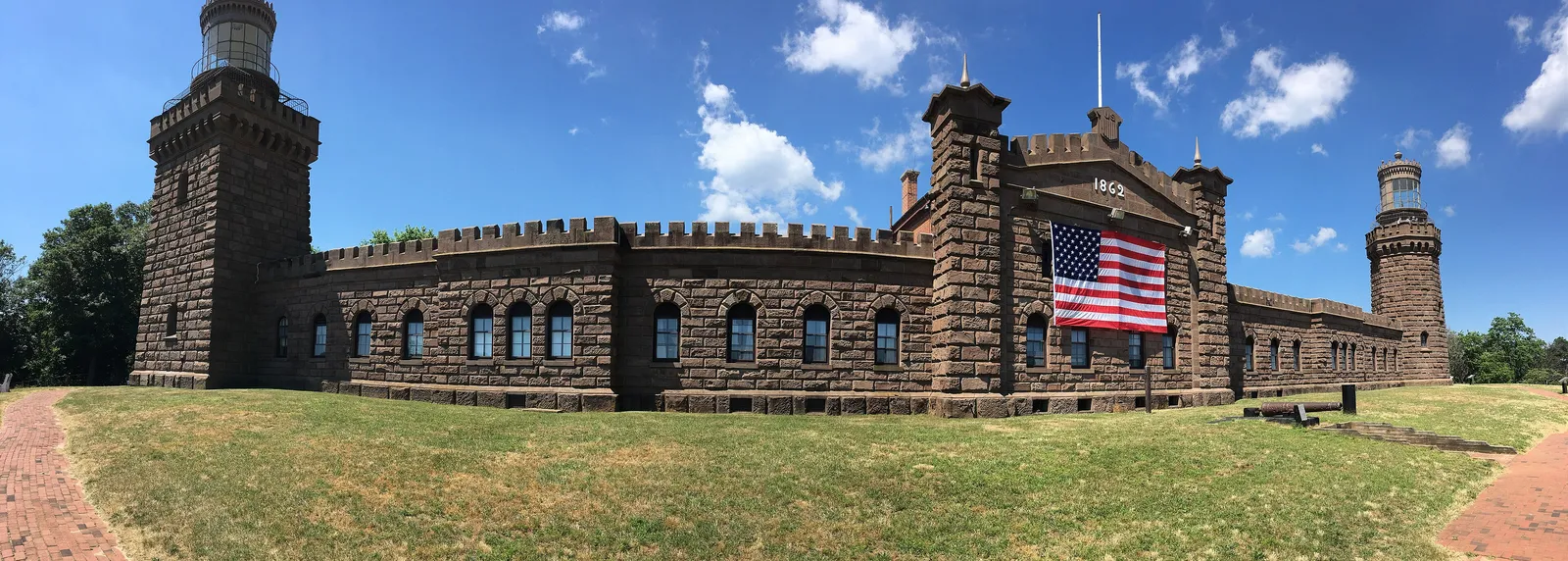 A long brownstone lighthouse building with a tower at each end and a large American flag over the entrance, behind a wide lawn