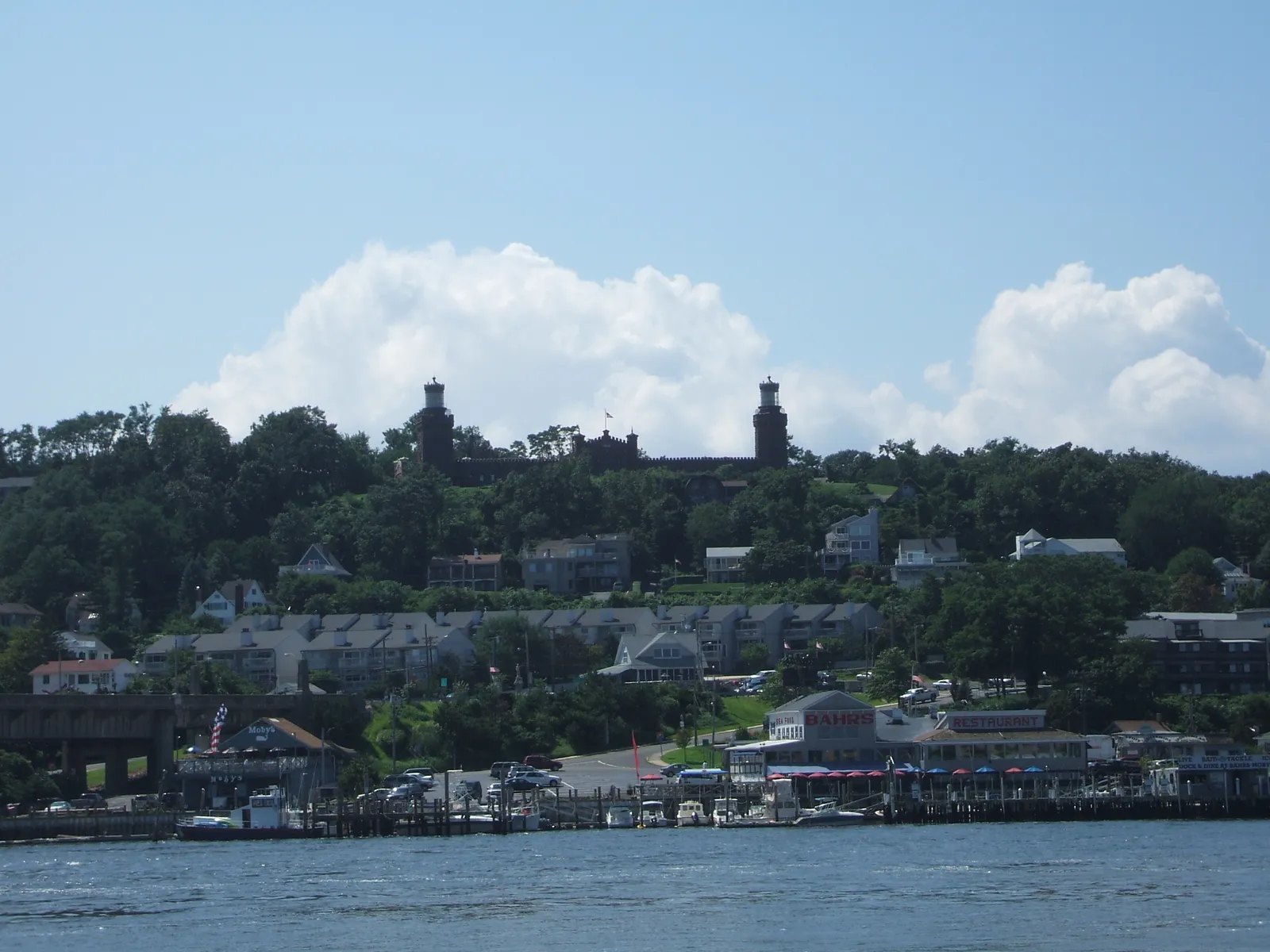 Two stone lighthouse towers on a wooded hilltop above waterfront homes and docks along the river