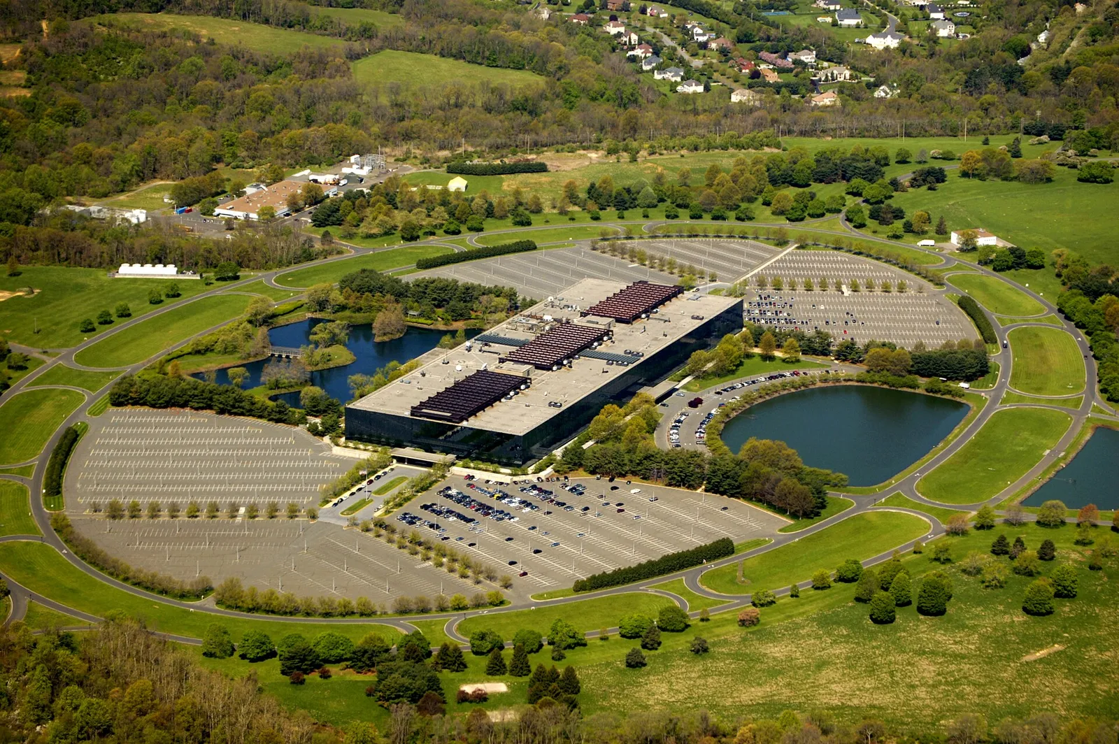 Aerial view of the mirrored Bell Labs building in Holmdel, now Bell Works, ringed by lakes, parking fields and an oval road through green lawns
