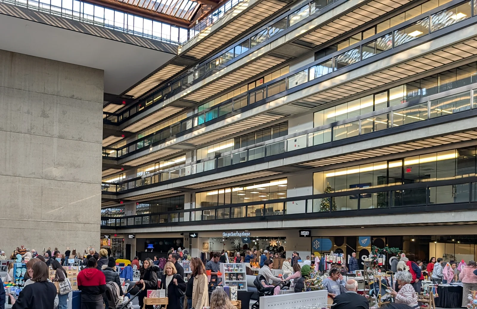 Shoppers crowding vendor tables at the indoor farmers market in the Bell Works atrium, with walkways and a skylight above
